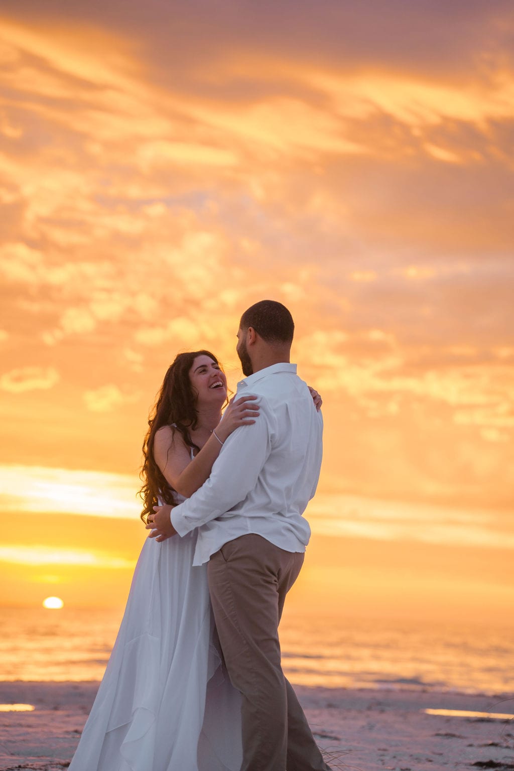 Couple laughing embrace fiery orange sunset beach engagement