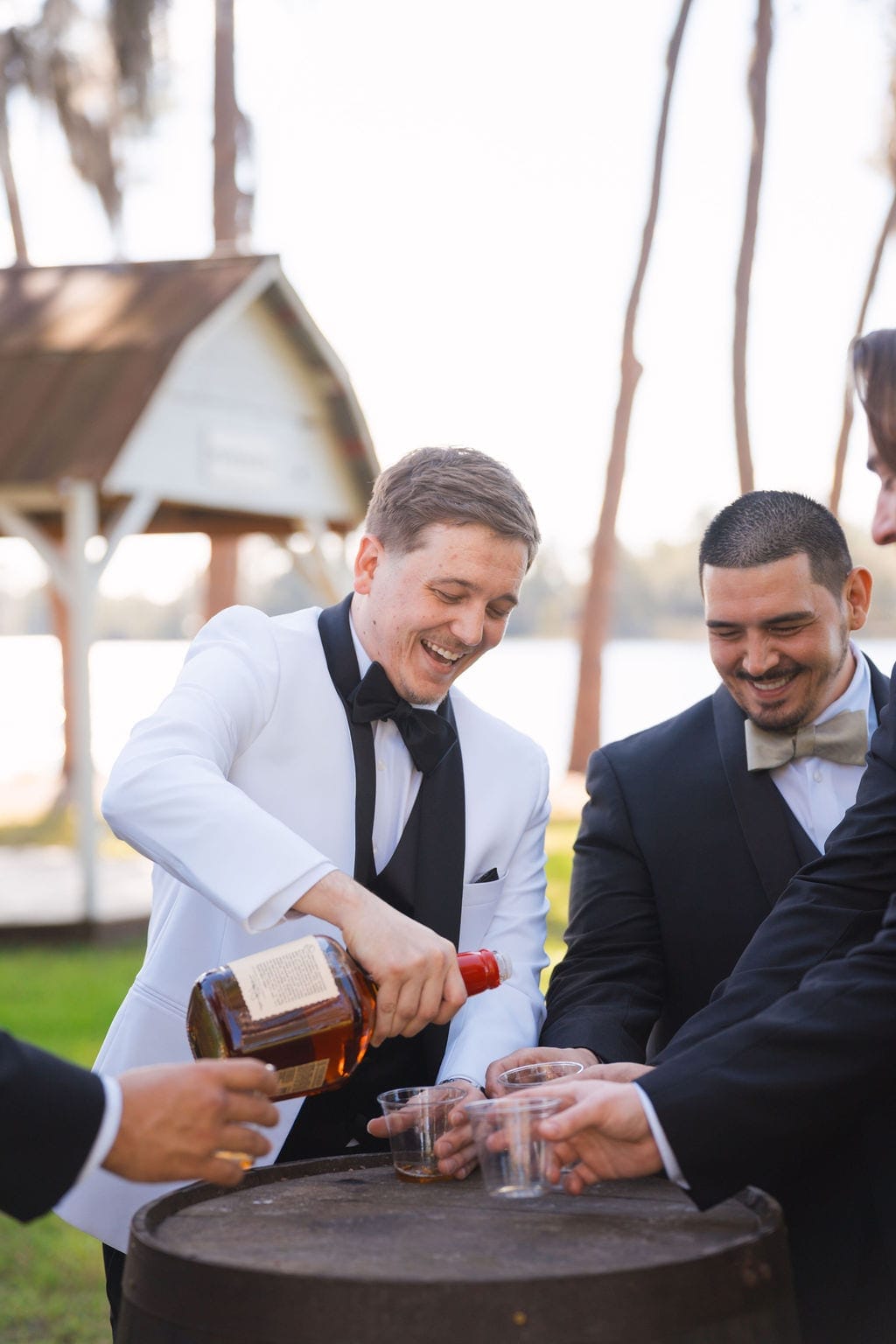 Groom pouring whiskey with groomsmen lakeside barrel toast
