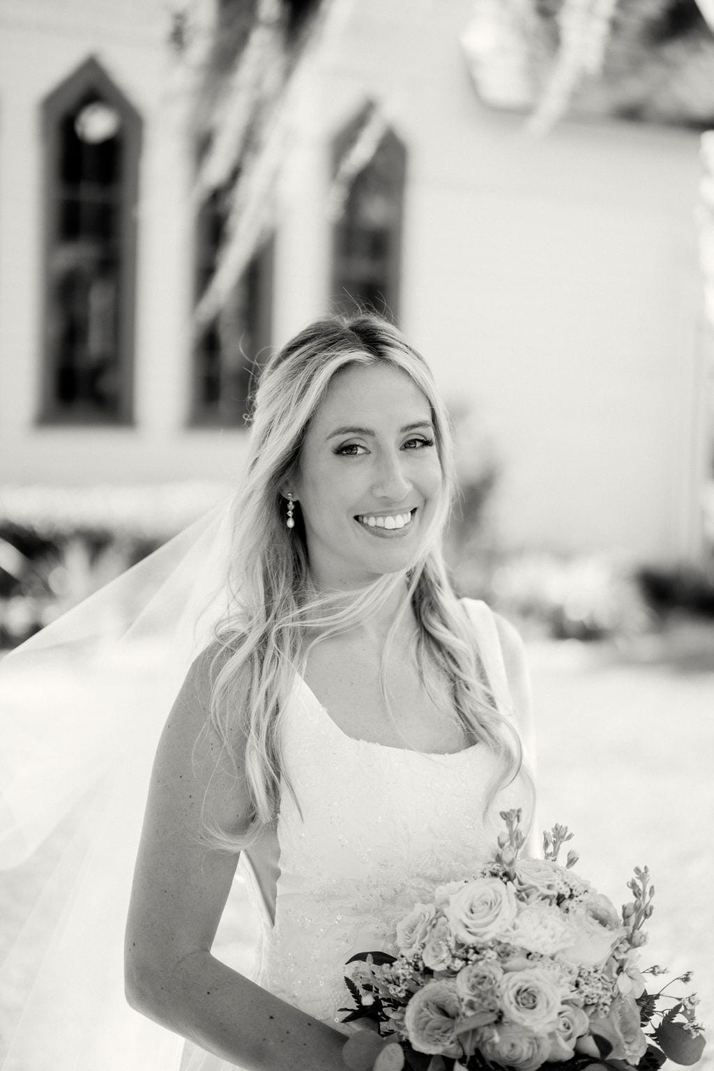 Bride smiling with veil and bouquet outside white chapel
