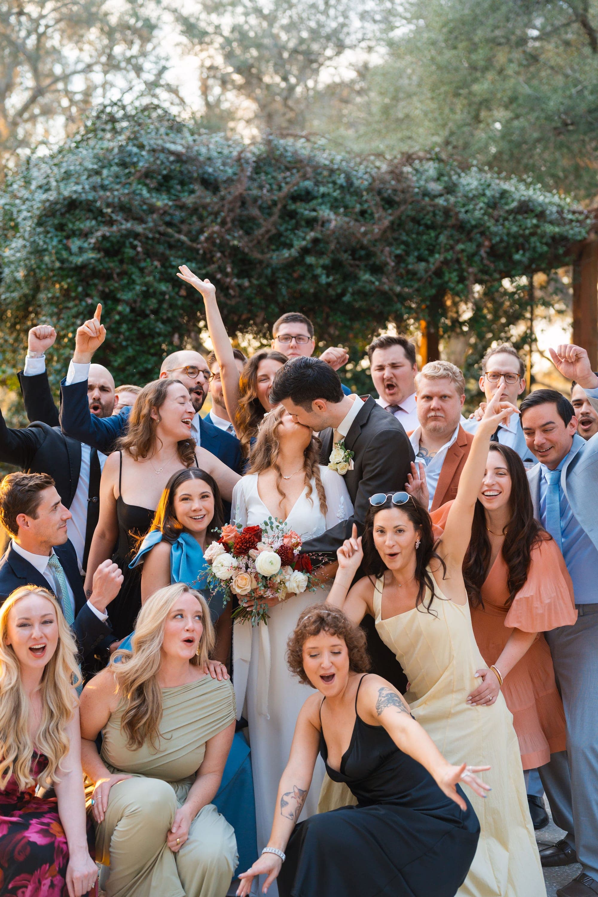 Newlyweds kissing while their wedding party cheers around them in a garden