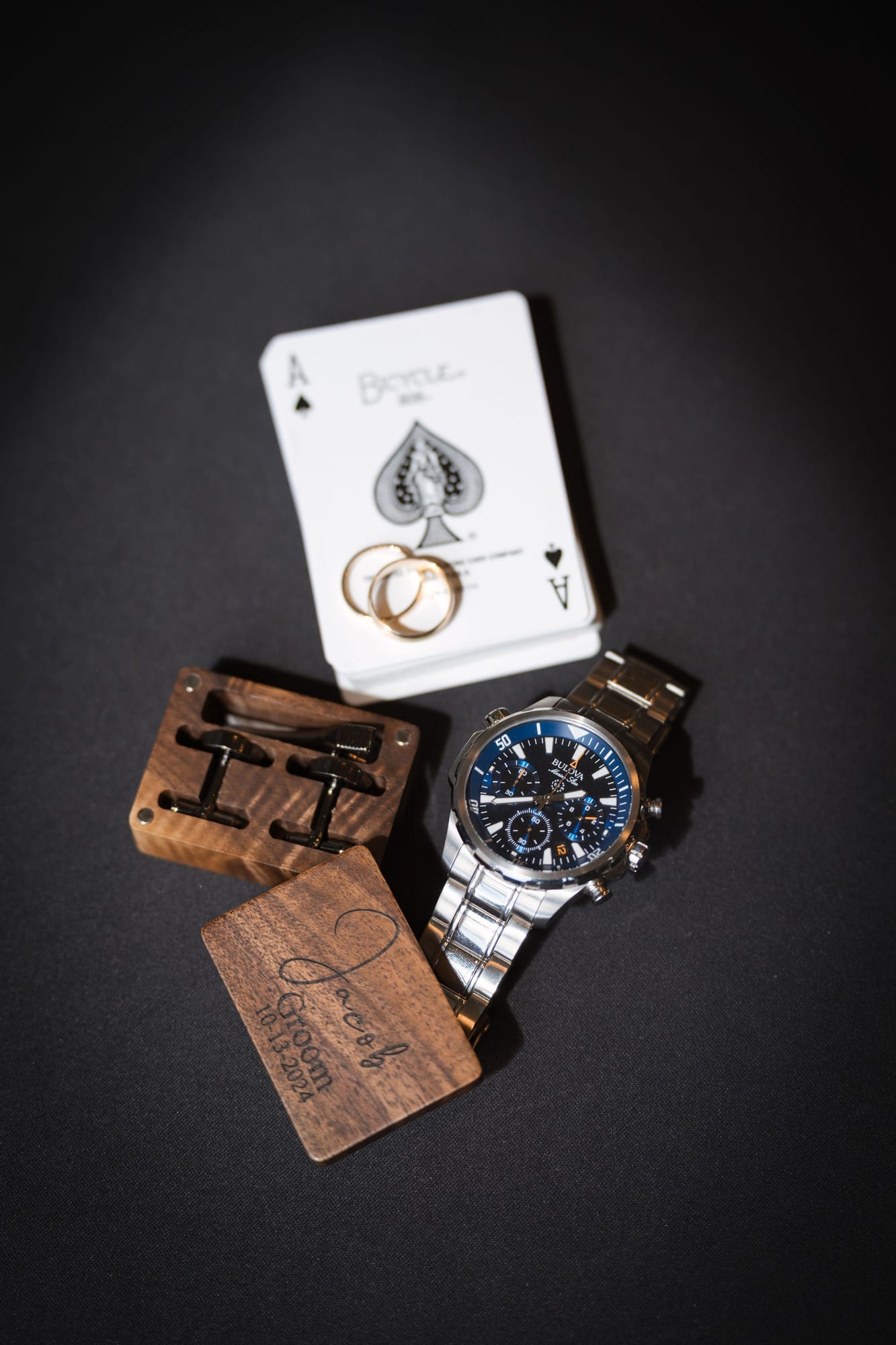 Groom's watch, wedding band, and an ace of spades card laid out on a dark box