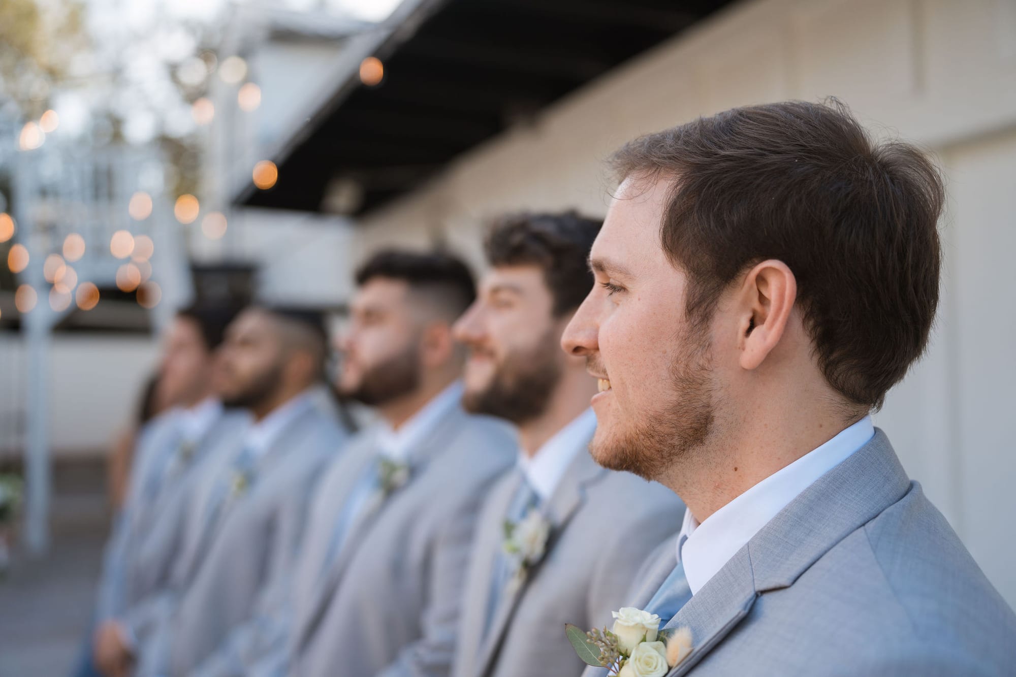Groomsmen in light gray suits lined up under string lights